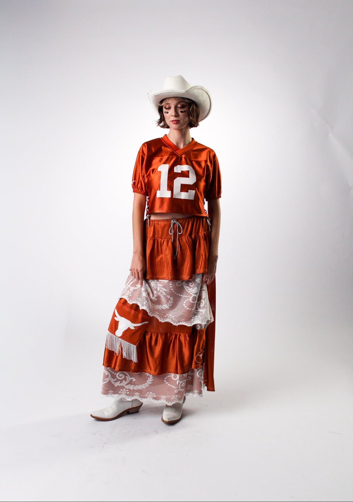 A model wearing a Texas-themed costume consisting of an orange short-sleeve jersey top with the number 12, a matching orange and white tiered skirt with a white lace overlay, a white cowboy hat, and white cowboy boots, standing against a white studio backdrop.