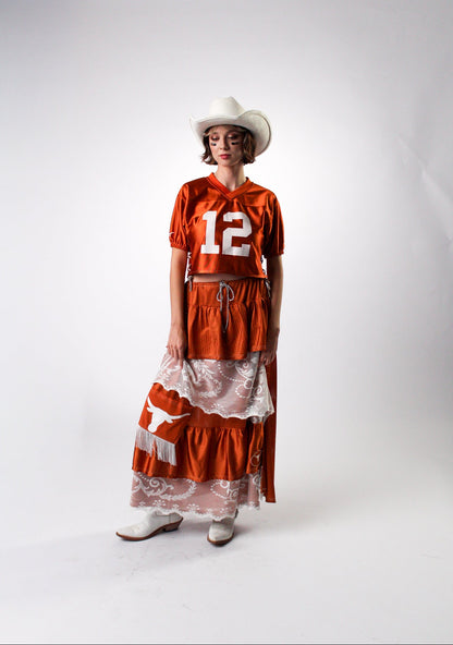 A model wearing a Texas-themed costume consisting of an orange short-sleeve jersey top with the number 12, a matching orange and white tiered skirt with a white lace overlay, a white cowboy hat, and white cowboy boots, standing against a white studio backdrop.